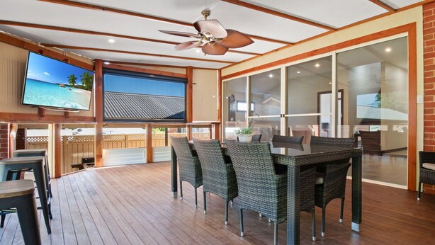 A covered outdoor dining area with wicker furniture, a ceiling fan, and a TV displaying a beach scene.