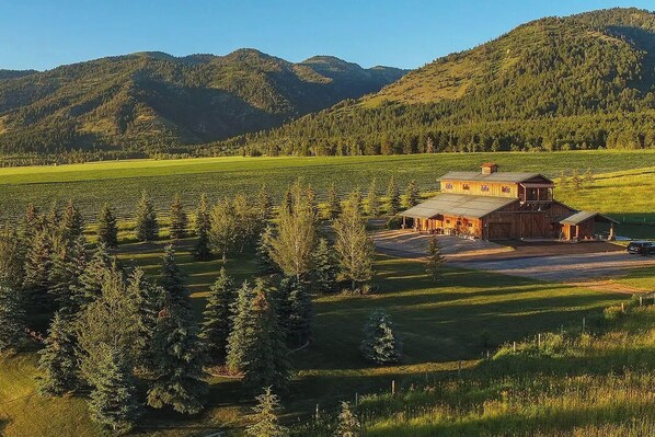 A rustic wooden barn with a large open area in front, surrounded by a forested landscape and mountains in the background.