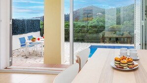 A dining area with a table set for six, a glass door leading to a balcony with a pool, and a scenic view of hills and trees.