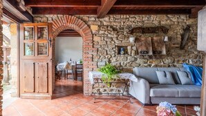 Ein Innenraum mit Steinwand, Holzbalkendecke und Kamin. Es gibt eine Essecke mit Tisch und Stühlen sowie eine Couch im Vordergrund.