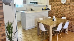Une cuisine avec des meubles blancs, une table à manger en bois avec des chaises blanches, une horloge au mur et une plante en pot.
