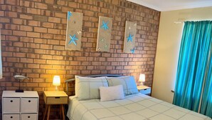 Une chambre avec un mur en briques, un lit recouvert d’une courtepointe matelassée, deux tables de chevet avec des lampes, une table de nuit, une commode et une fenêtre avec des rideaux.