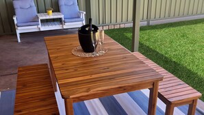 Une table de patio en bois, accompagnée de deux bancs, d’une plante en pot et d’une coupe de champagne.
