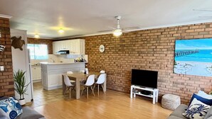 Un salon avec deux canapés gris, une table à manger avec quatre chaises, un téléviseur posé sur un meuble et un mur en briques.