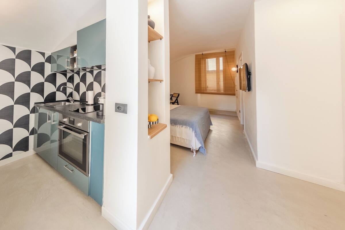 A compact kitchen with modern appliances and a geometric backsplash, leading to a bedroom with a bed and a bedside table.