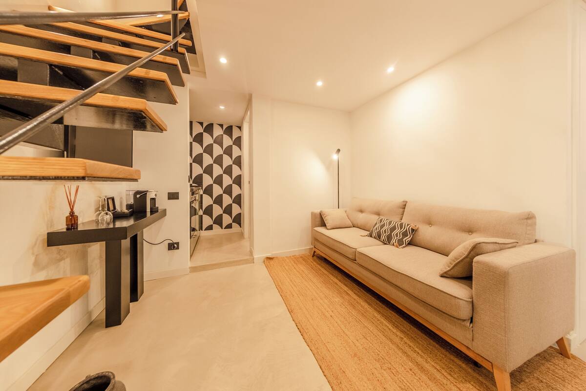 A modern living room with a beige sofa, a wooden staircase, and a patterned wall.