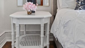 Un dormitorio con una cama blanca, una mesita de noche con lámpara, un espejo y un cojín estampado.