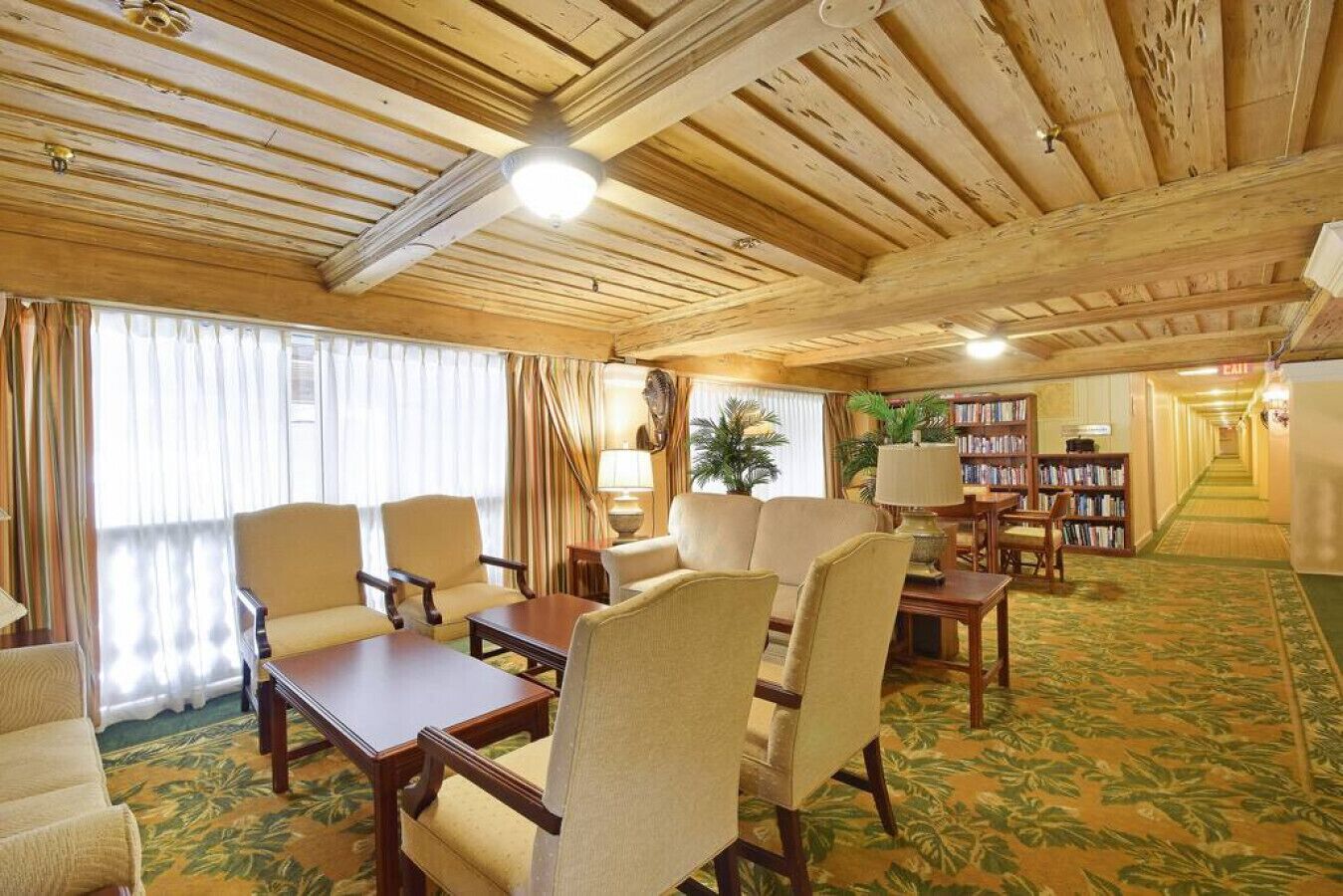 A spacious room with wooden ceilings, a large window with curtains, a sofa, chairs, a table, and a bookshelf.