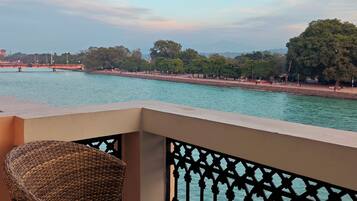 A balcony with a wicker chair overlooking a river, with trees and a distant bridge in the background.