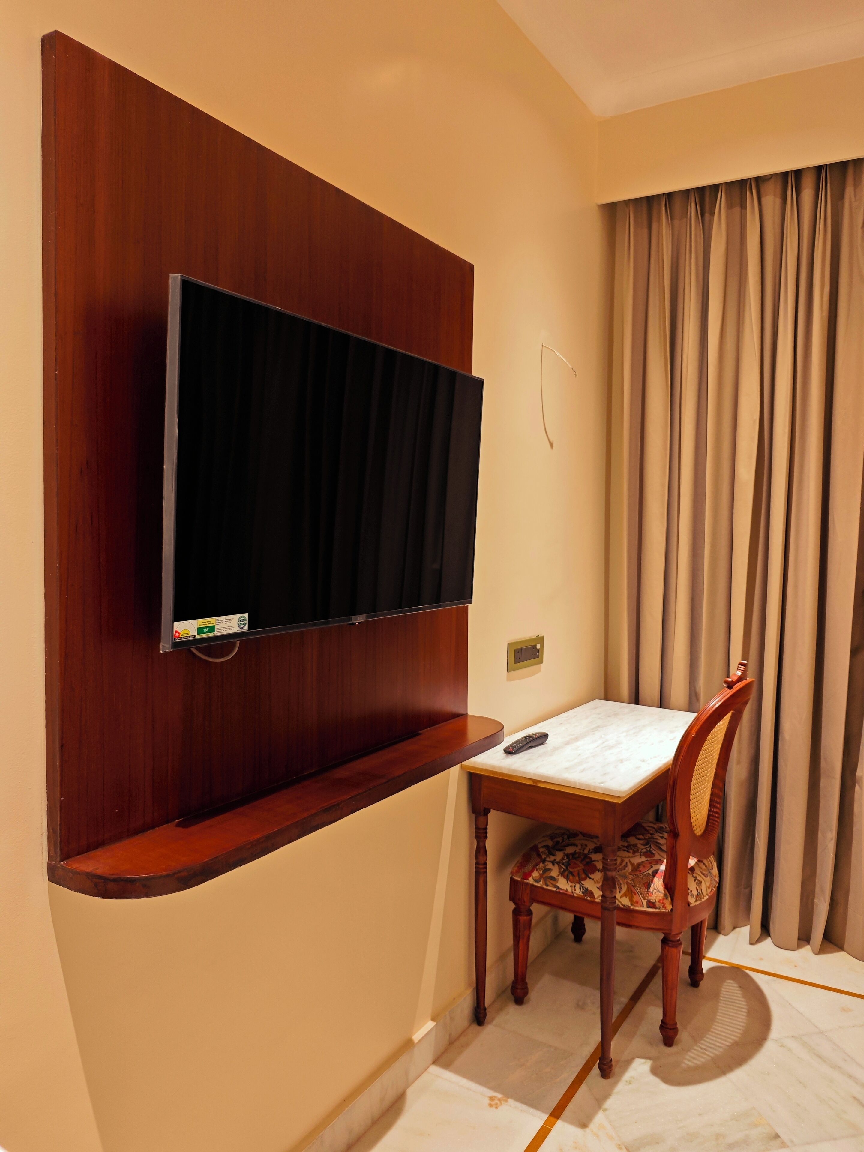Une chambre d’hôtel dotée d’un téléviseur à écran plat fixé sur un panneau en bois, d’une petite table avec une télécommande, d’une chaise en bois avec un coussin à motifs et d’une grande fenêtre avec des rideaux.