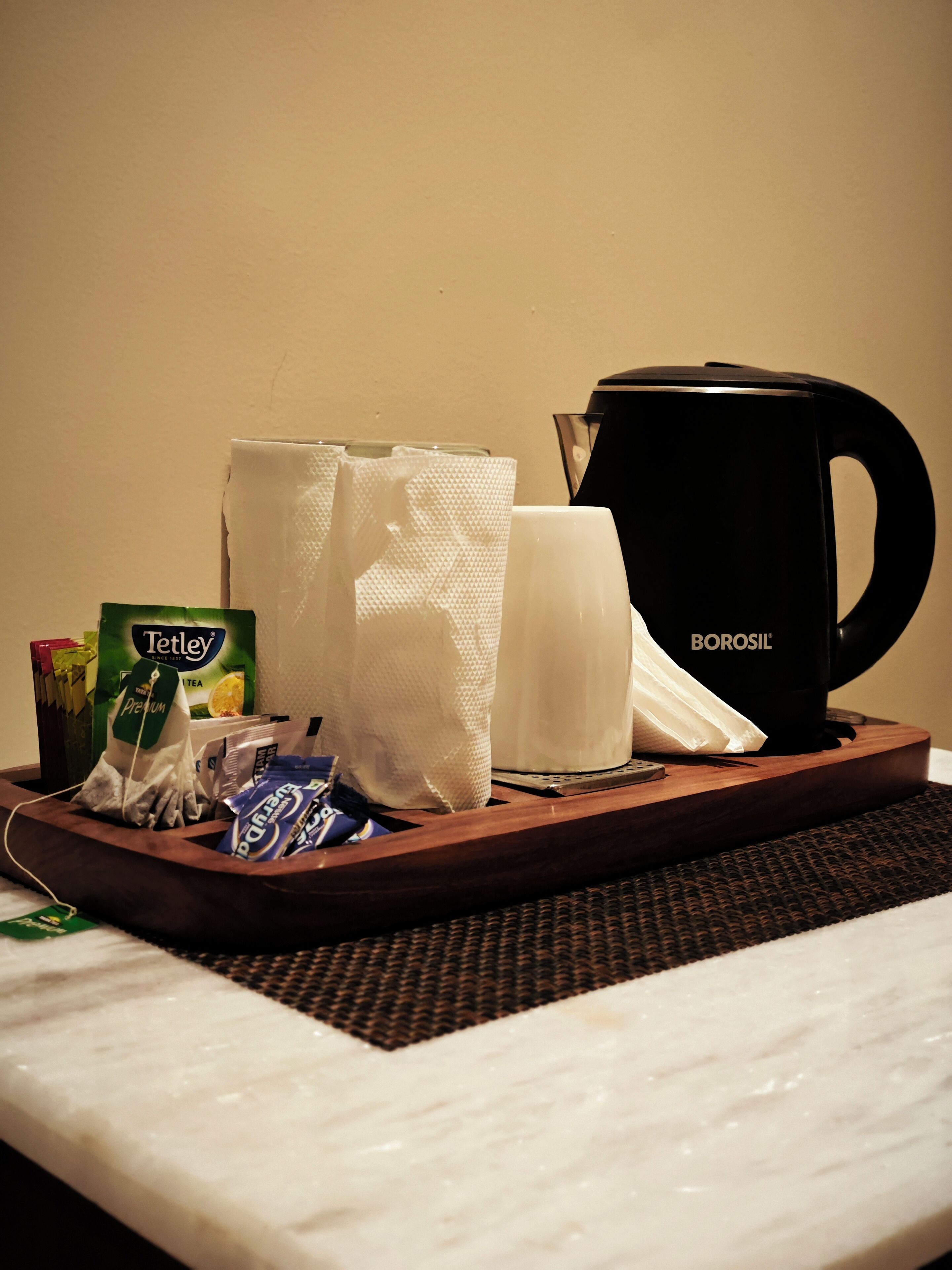 A tray with a black coffee maker, a tissue box, a tissue roll, a tea bag, and a packet of sugar on a bed.