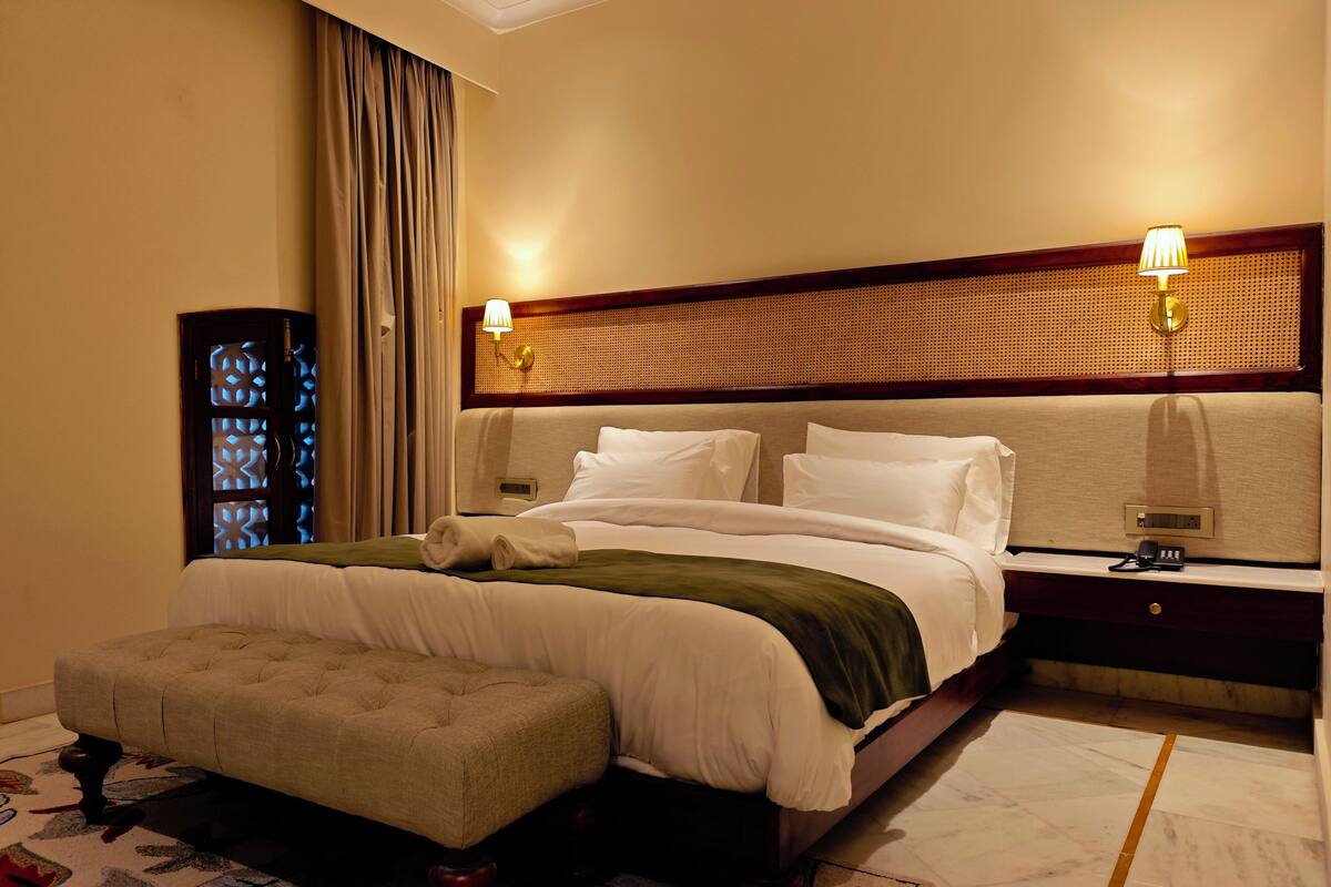 A hotel room with a large bed, a nightstand, a lamp, a window with curtains, and a patterned rug.