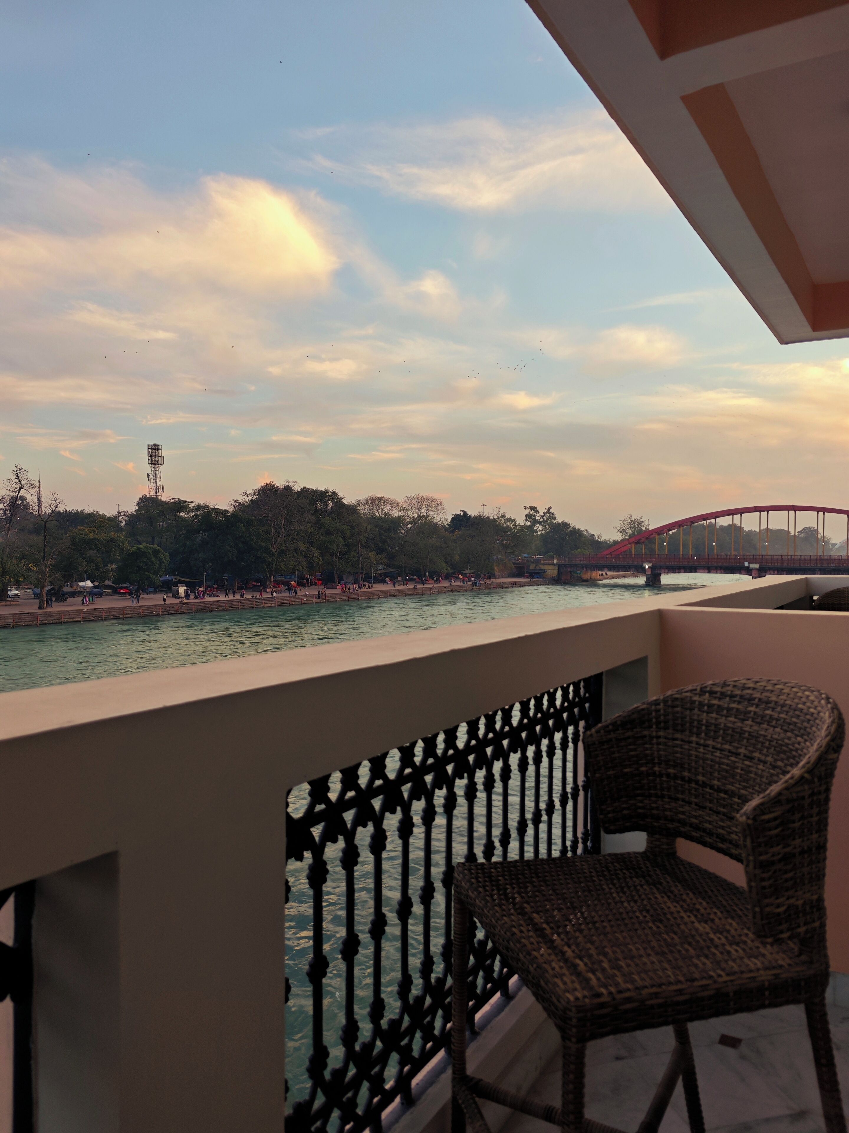 A balcony with a wicker chair, overlooking a river with a bridge and trees in the background.