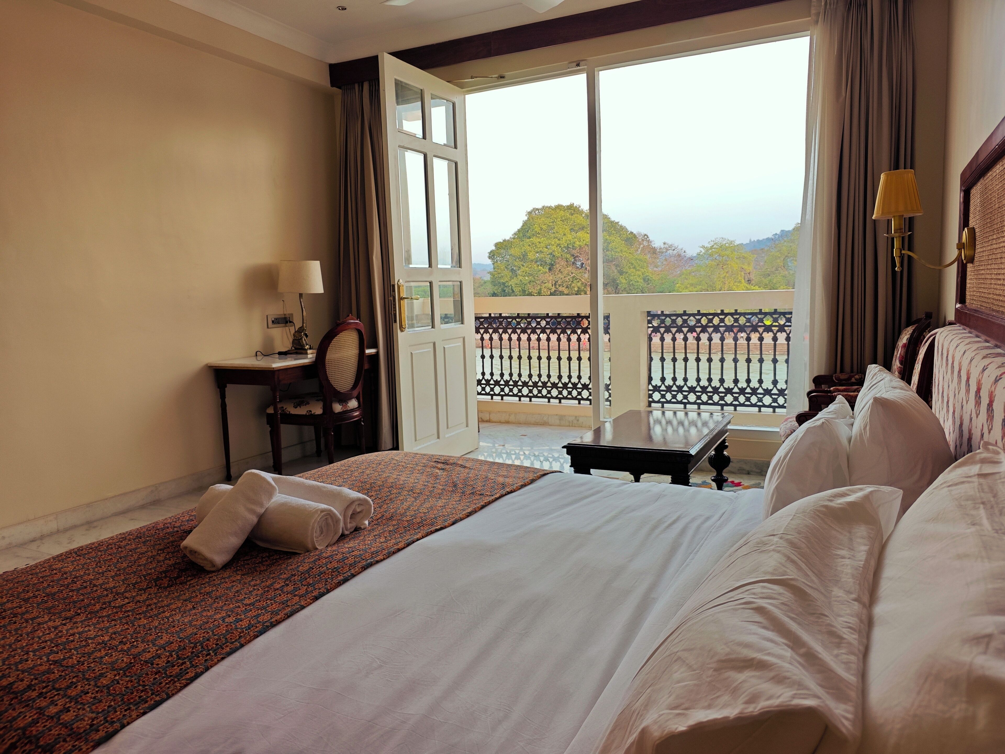 Quarto de hotel com uma cama grande, uma escrivaninha com abajur e vista para uma varanda com guarda-corpo.