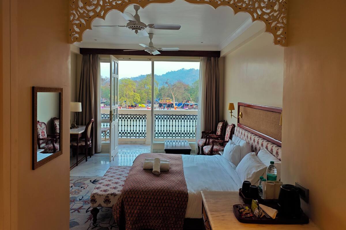 A hotel room with a bed, a desk, a chair, a balcony with a railing, and a view of the outdoors.