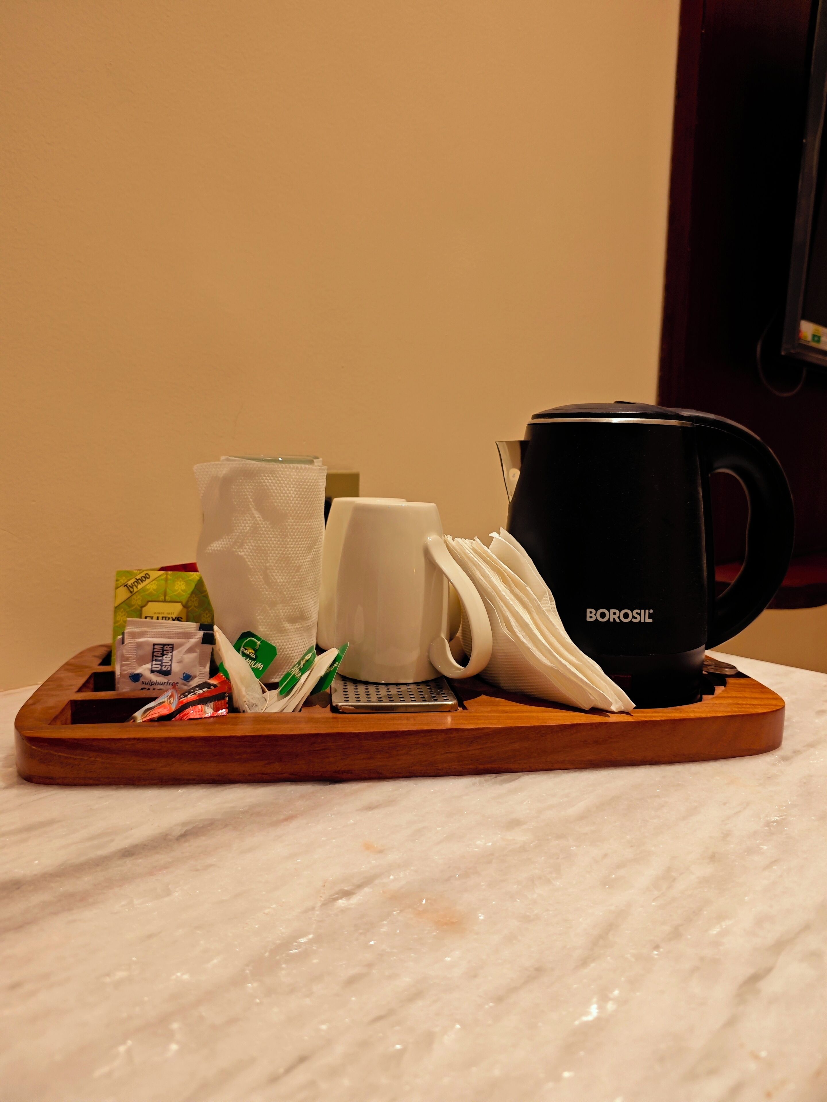 Uma bandeja com uma cafeteira, uma caneca branca, um estojo de lenços e alguns artigos de higiene pessoal sobre uma superfície de mármore.