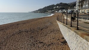 A coastal beach with a stone wall and railings, a clear sky, and buildings in the background.