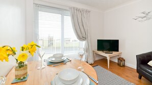 A dining area with a table set for two, a vase of yellow flowers, a television, and a black leather couch.