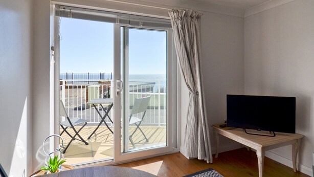 A room with a sliding glass door leading to a balcony with a view of the sea. There is a TV on a wooden stand and a small table with a plant.