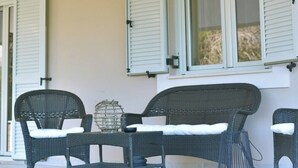 Un espace extérieur aménagé avec des meubles en osier et une table, un vase décoratif en métal et une fenêtre avec des volets.