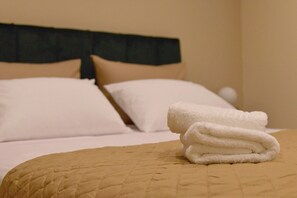 Een net opgemaakt bed met een beige sprei en witte kussens, een opgevouwen witte handdoek op het bed, en een donker hoofdbord.