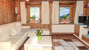 Een knus kamer in chaletstijl met een bank, een bijzettafeltje en een televisie. Er zijn ramen met gordijnen, een radiator en een plant op de vensterbank.