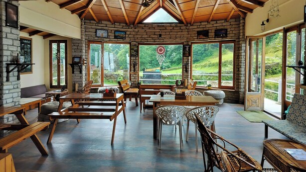 A spacious dining area with wooden tables and chairs, large windows offering a view of the outdoors, and a stone wall with artwork.