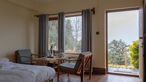 A hotel room with a bed, two chairs, a table, and a view of a garden through a large window.