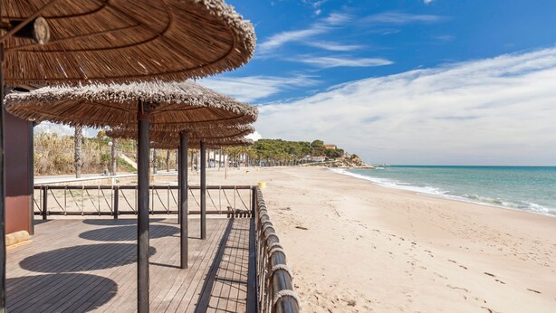 Een strand met houten terras en rieten parasols, een zandstrand en een gebouw op de achtergrond.