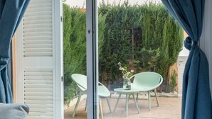 Een kamer met een schuifglazen deur die uitkomt op een terras buiten, met twee groene stoelen en een kleine tafel met een bloevas.