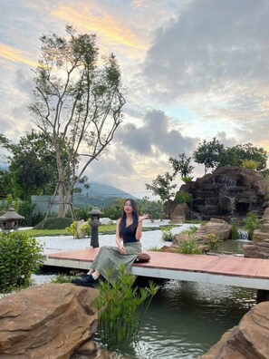 A woman sitting on a wooden deck by a pond with a waterfall and trees in the background.