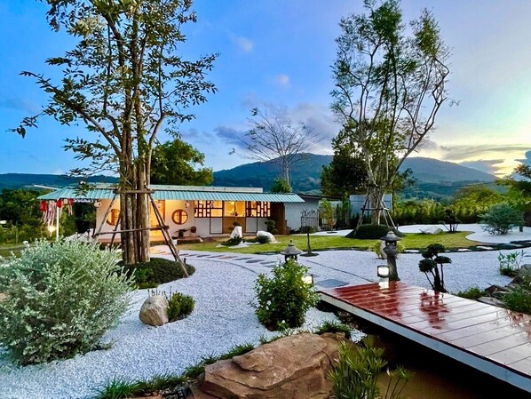 A traditional Japanese-style building with a wooden deck, surrounded by a landscaped garden with rocks and trees.