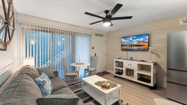 A modern living room with a sofa, a coffee table, a TV, a ceiling fan, and a kitchen area.