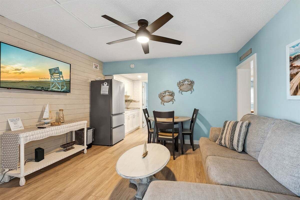 A living room with a sofa, a dining table, a TV, a ceiling fan, and a kitchen in the background.