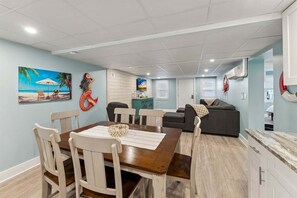 A dining area with a wooden table and chairs, a painting of a beach scene, a sofa, and a kitchen area with cabinets and a countertop.