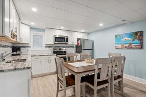A modern kitchen with white cabinets, a microwave, stove, and refrigerator. There is a dining table with chairs and a beach-themed painting on the wall.
