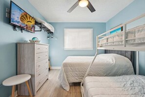 Une chambre avec un lit superposé, un téléviseur fixé au mur, une commode et une fenêtre avec des stores.