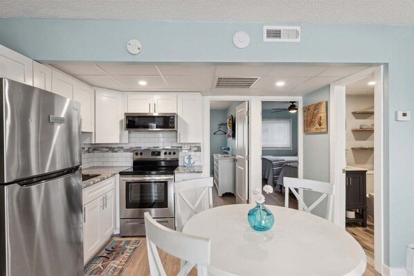 A compact kitchen with white cabinets, a stainless steel refrigerator, a microwave, and an oven. There is a dining table with chairs and a vase with flowers.