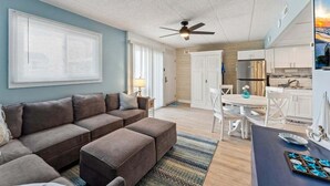 Un salon avec un canapé, une table à manger avec des chaises, un coin cuisine avec des placards et des appareils électroménagers, et un ventilateur de plafond.