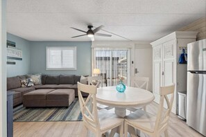 A living room with a sectional sofa, a dining table with chairs, a ceiling fan, and a kitchen area with a refrigerator.