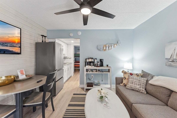 A modern living room with a sofa, coffee table, and a dining area with chairs and a table. There is a ceiling fan, a wall-mounted shelf with books, and a framed picture on the wall.