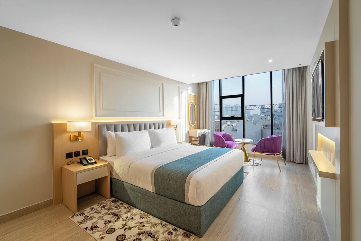 A modern hotel room with a large bed, a desk with a phone, a chair, and a view of the city.