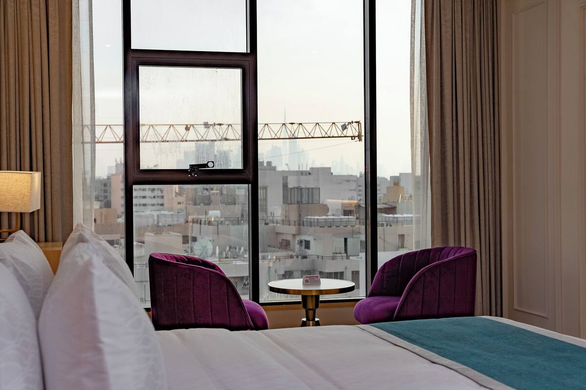A hotel room with a large window, a bed, two purple chairs, a small round table, and a lamp.