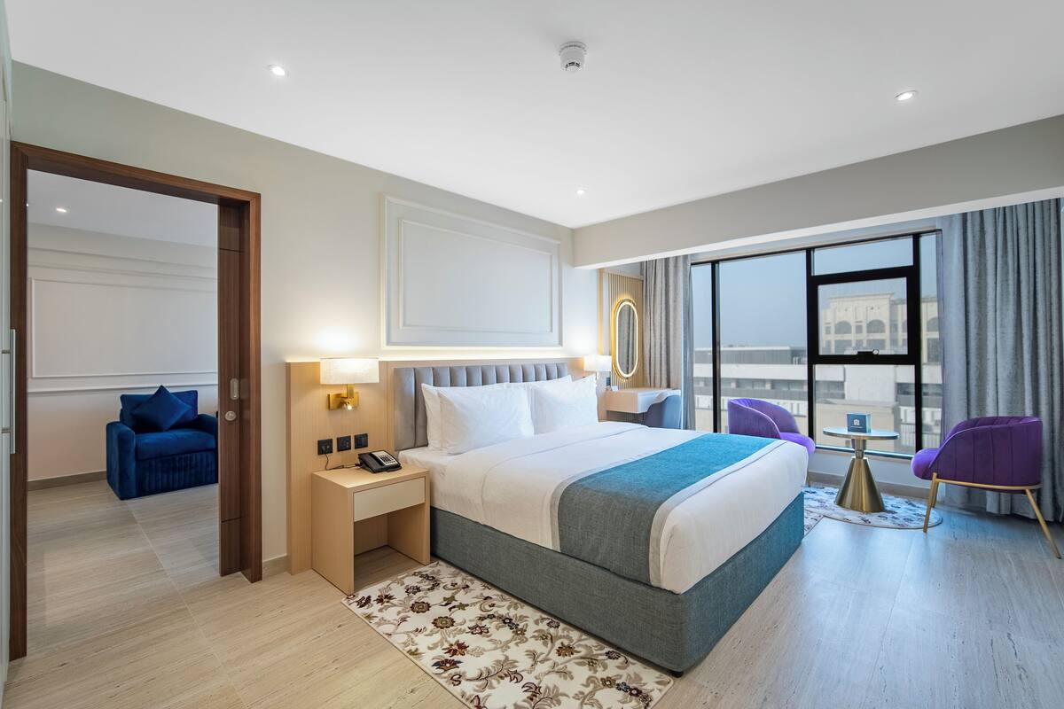A modern hotel room with a large bed, a sitting area with a small table and chairs, and a view of a cityscape through large windows.