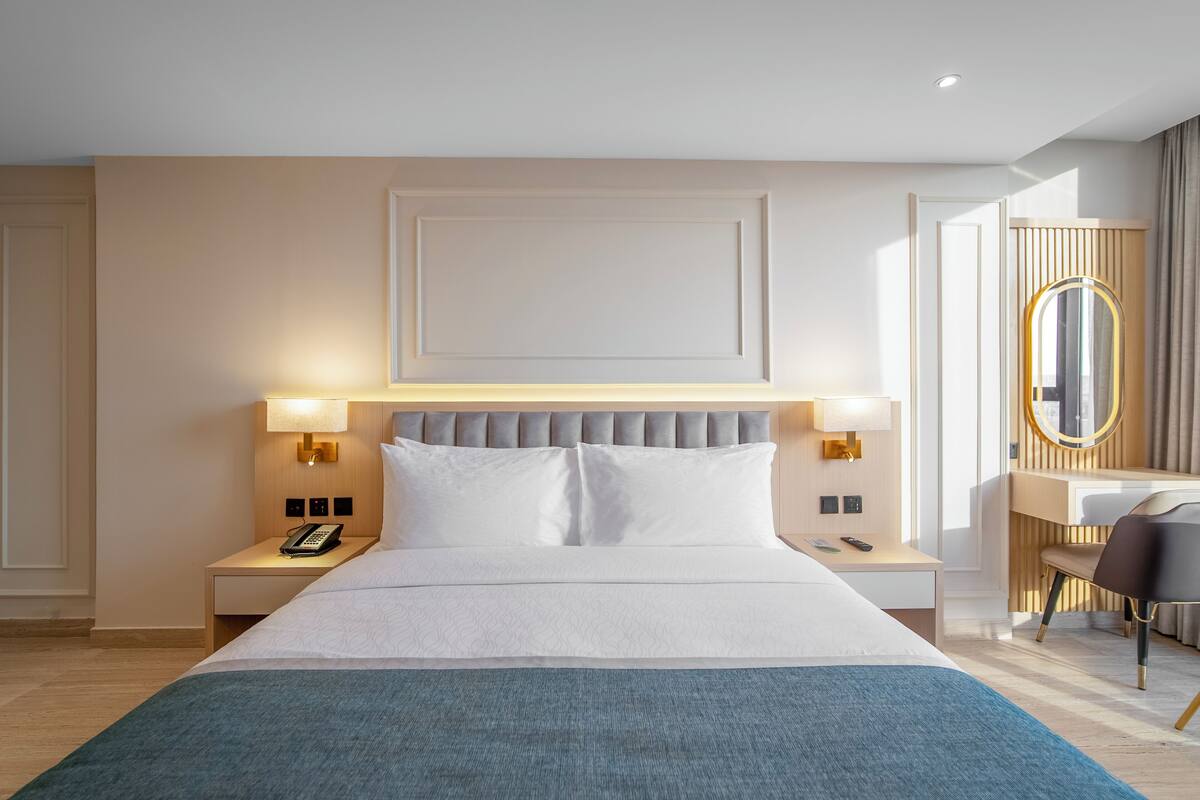 A modern hotel room with a large bed, bedside lamps, a desk with a chair, and a mirror.