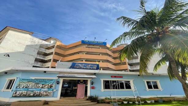A multi-story building with a blue facade, a sign for 'Apartment Sewa Bulan' indicating monthly rental, and a palm tree in the foreground.