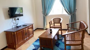 A room with a TV mounted on the wall, a wooden cabinet, a table with chairs, and a window with curtains.