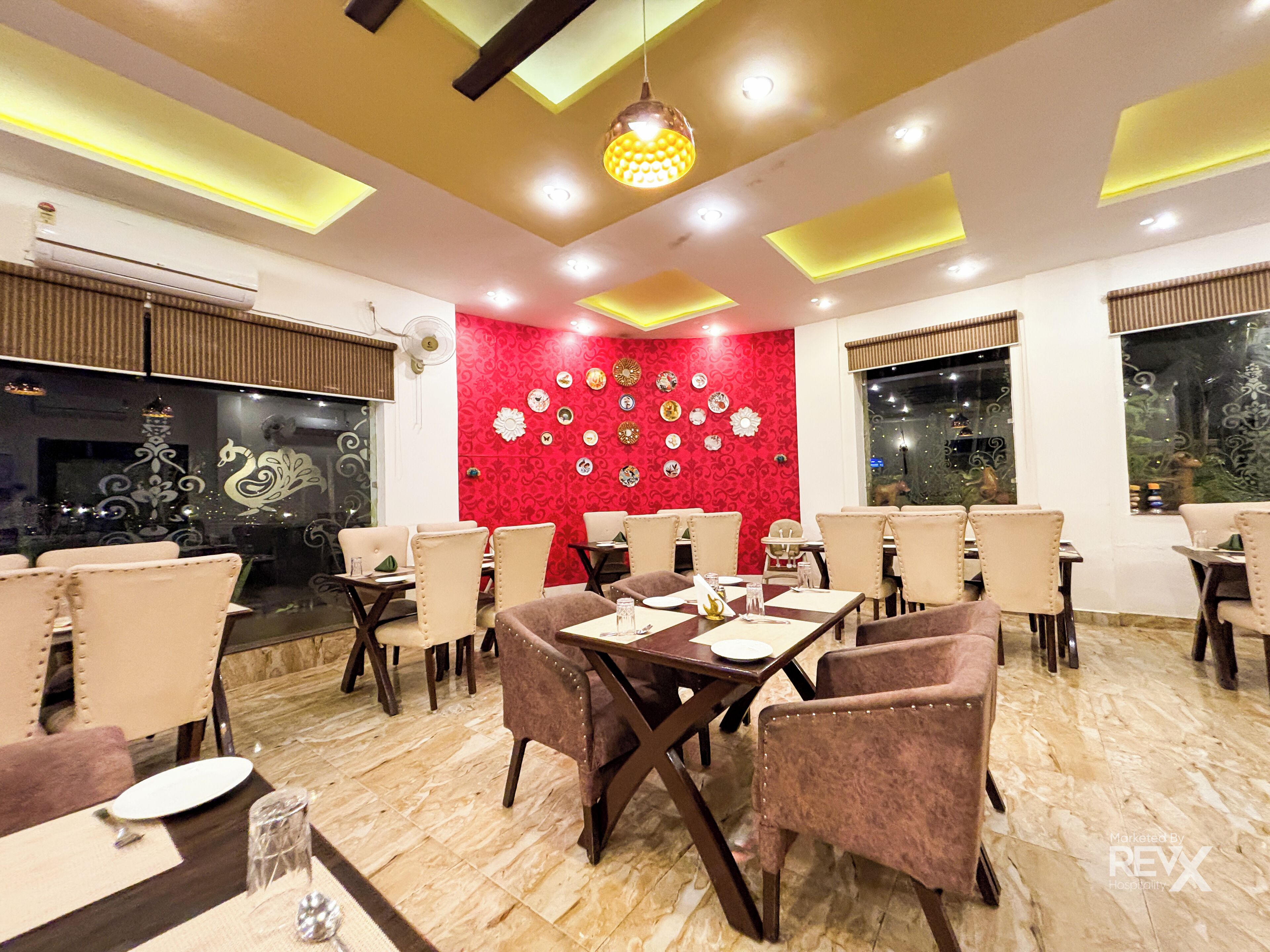 A restaurant with a red wall decorated with plates, beige upholstered chairs, and a modern ceiling design.