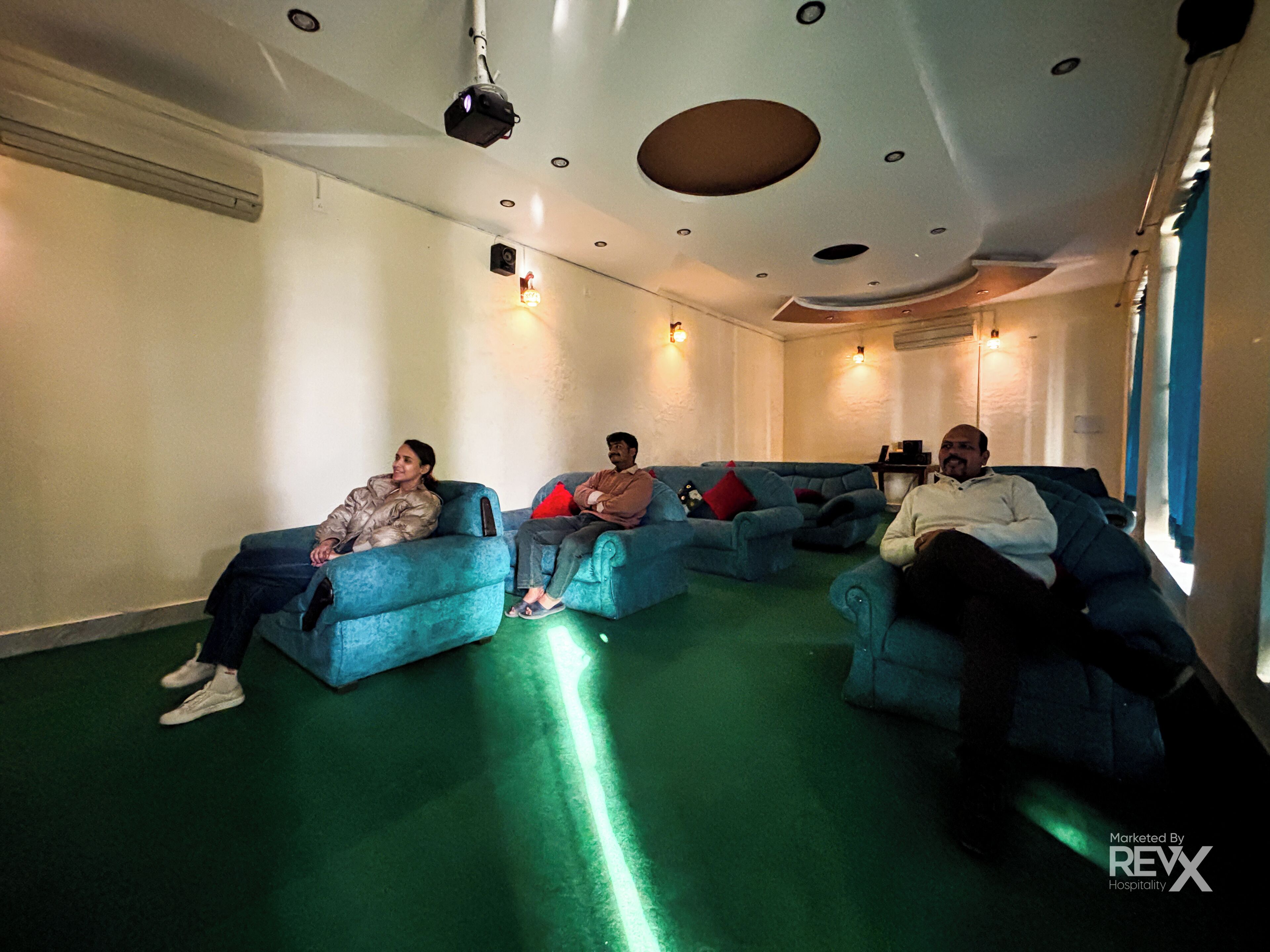 A room with blue sofas arranged in a semi-circle, a ceiling-mounted projector, and a green carpet.