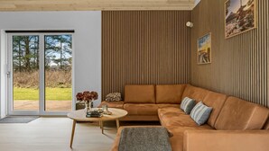 Una sala moderna con un sofá grande de cuero marrón, una mesa redonda de centro y vista a un área exterior con césped a través de una puerta de vidrio.